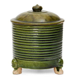 A rare Chinese green-glazed pottery tripod jar and cover, Tang dynasty, the cylindrical ribbed body is covered in a brilliant green glaze which pools between t