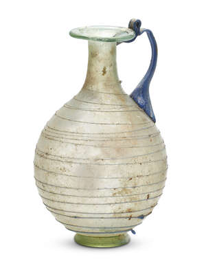 A small Roman light green glass jug, 1st century A.D., the flaring mouth with an infolded rim on a pear-shaped body and hollow tubular base-ring, decorated wit