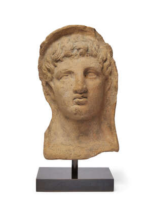 An Etruscan terracotta votive head of a young man, circa 3rd-2nd Century B.C., of hollow form, wearing a himation drawn over wavy hair, with almond shaped lidd