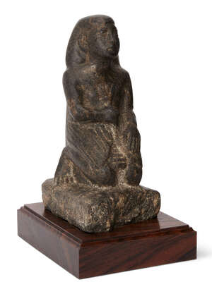 A reproduction granite Egyptian style kneeling figure wearing a shendyt kilt and bag wig, the left hand on the knee, the right hand held to the chest and clasp