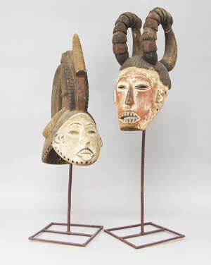 
	
		Two tribal hardwood masks, possibly Igbo, one with curving horns, his face with slit eyes...